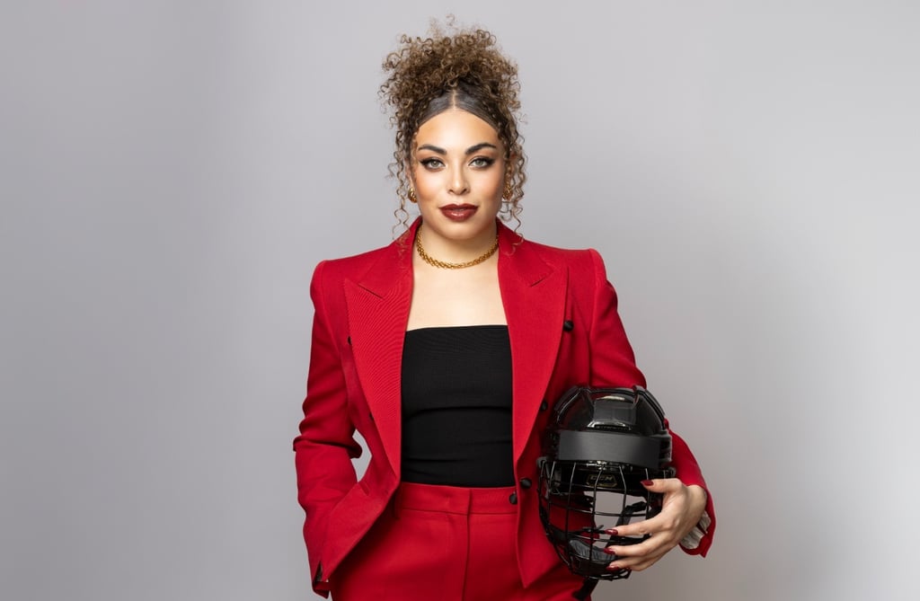 A woman in a red power suit posing with a black hockey helmet against a grey background.