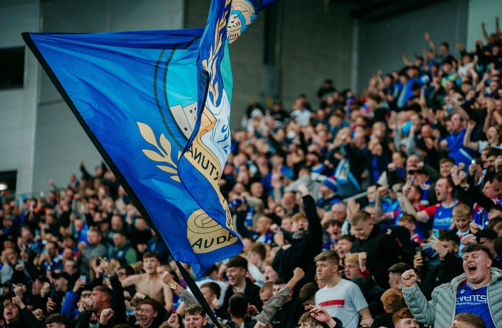 Linfield-Fan in blauem Trikot vor dem Windsor Park in Belfast, mit Blick auf die moderne Stadionfassade und die Tribünen im H