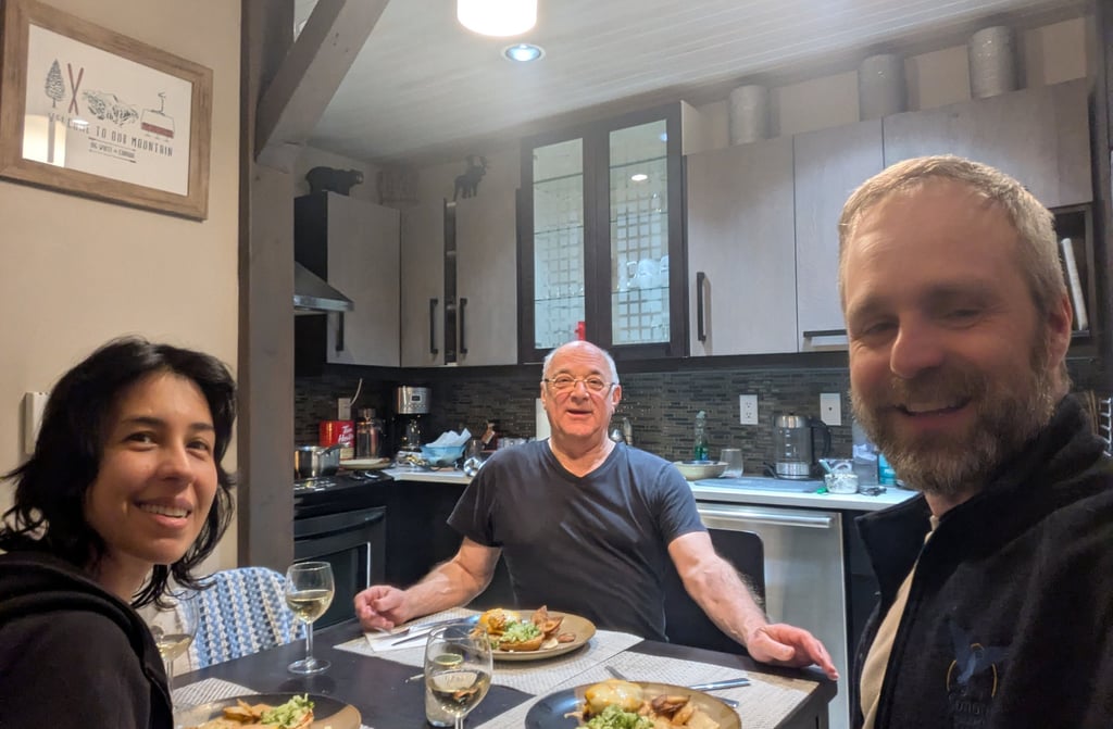 Three people sitting at a dining table in a kitchen, smiling while sharing a home-cooked meal.