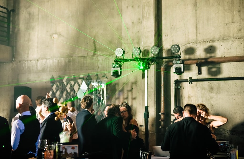 Guests dancing at a wedding reception with green laser light show and industrial concrete wall backdrop.