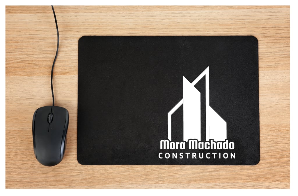 Black mouse pad with white Mora Machado Construction logo next to a computer mouse on a wood desk.