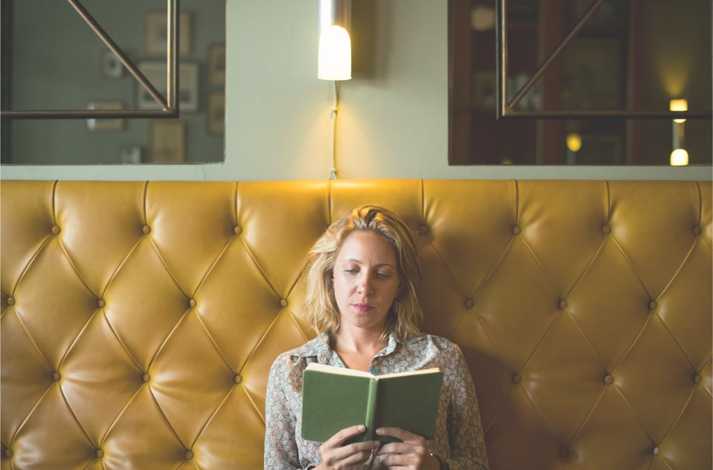 A blonde woman reading a book while sitting against a yellow tufted leather banquette in a cafe.