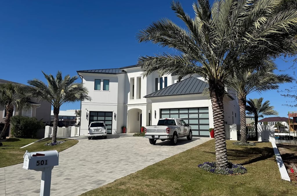 Modern white luxury home with a paved driveway, palm trees, and glass garage doors under a clear blue sky.