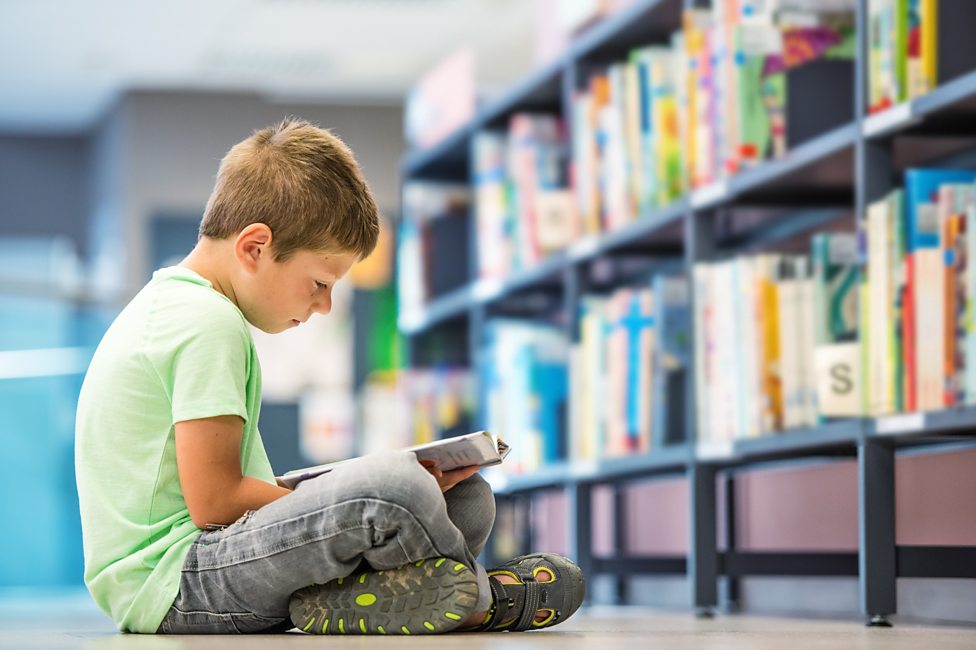imagem mostra uma criança de 10 anos aproximadamente lendo um livro sentado no chão da biblioteca