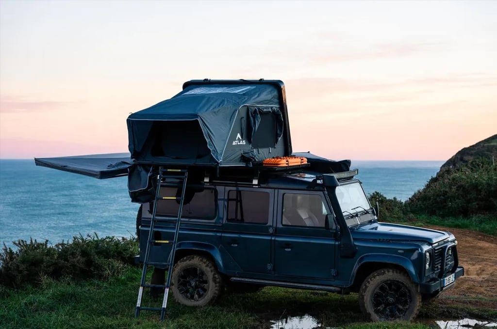 Land Rover Defender with a rooftop tent and awning parked on a scenic coastal cliff at sunset.