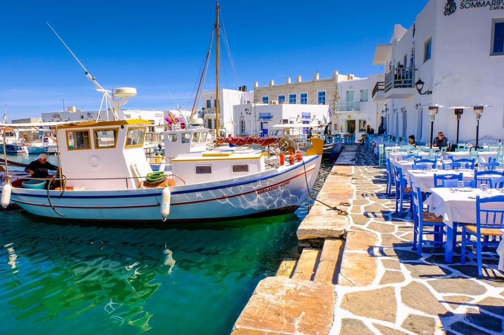 Traditional fishing boats docked by a waterfront Greek restaurant with blue chairs in Paros, Greece.