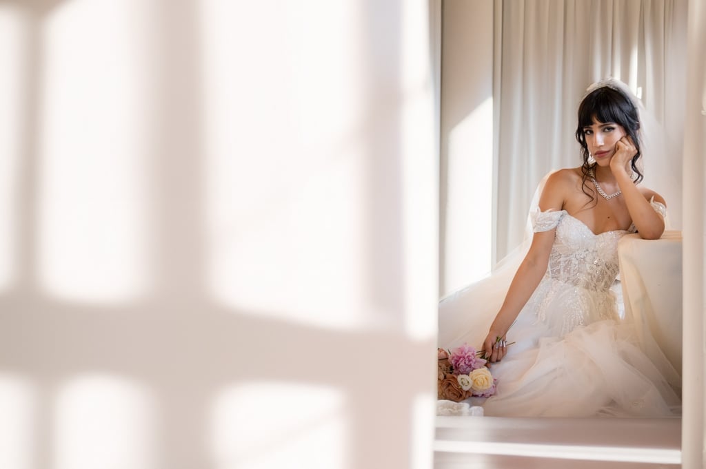 Sposa seduta in una stanza bianca, circondata da ombre geometriche di luce, con bouquet tra le mani