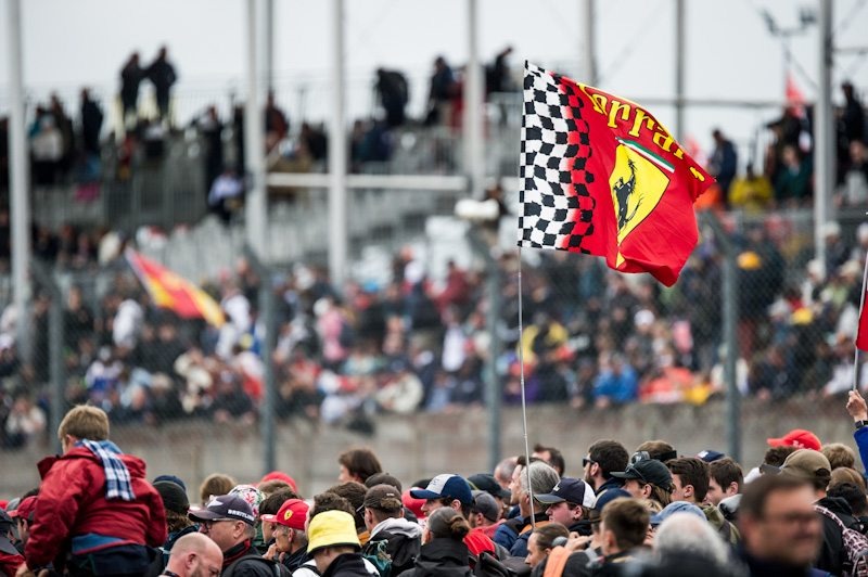 Ferrari flag waving over a crowded grandstand during a Formula 1 racing event at a motorsports track.