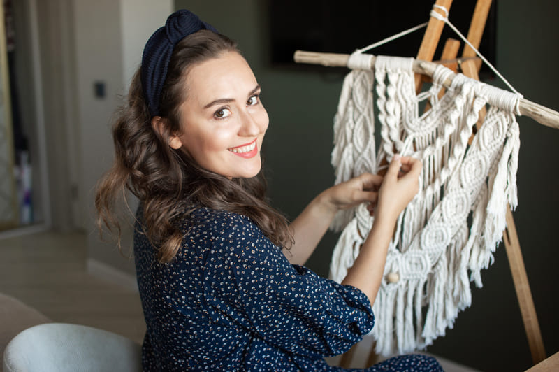 Mujer sonriente creando macramé hecho a mano en caballete de madera dentro de su estudio artesanal.