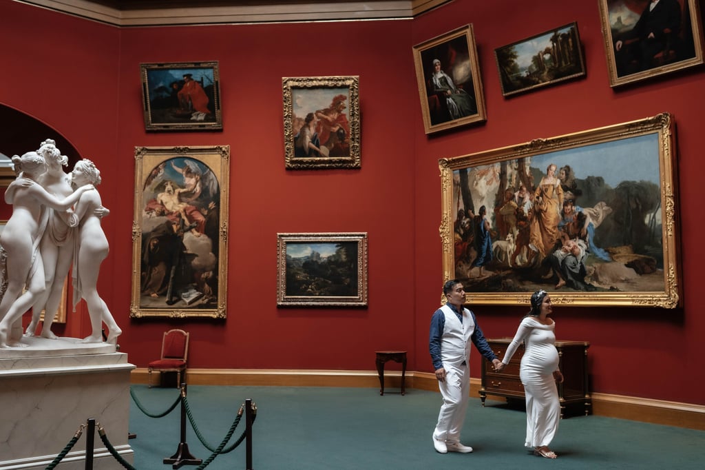 A pregnant couple holding hands walks through an art gallery with red walls and classical oil paintings.
