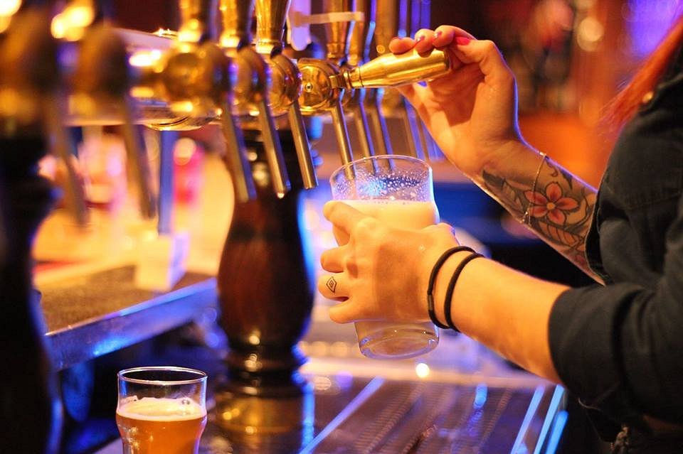 A bartender with tattoos pouring a fresh craft beer from a brass tap into a glass at a pub.