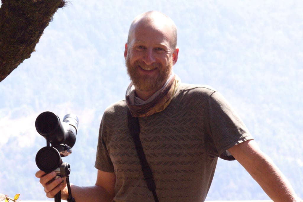 San Cristobal bird guide Benedict Simmons smiling with telescope, with binoculars in front hillside 