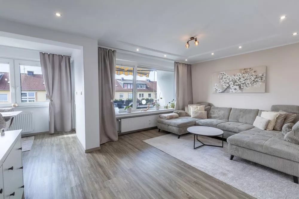 Modernes Wohnzimmer mit grauem Ecksofa, Holzboden und großen Fenstern mit neutralen Vorhängen.