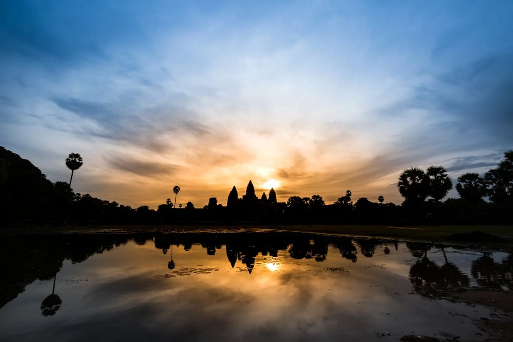 Angkor Wat temple silhouette at sunrise with dramatic orange sky and palm trees reflected in still w