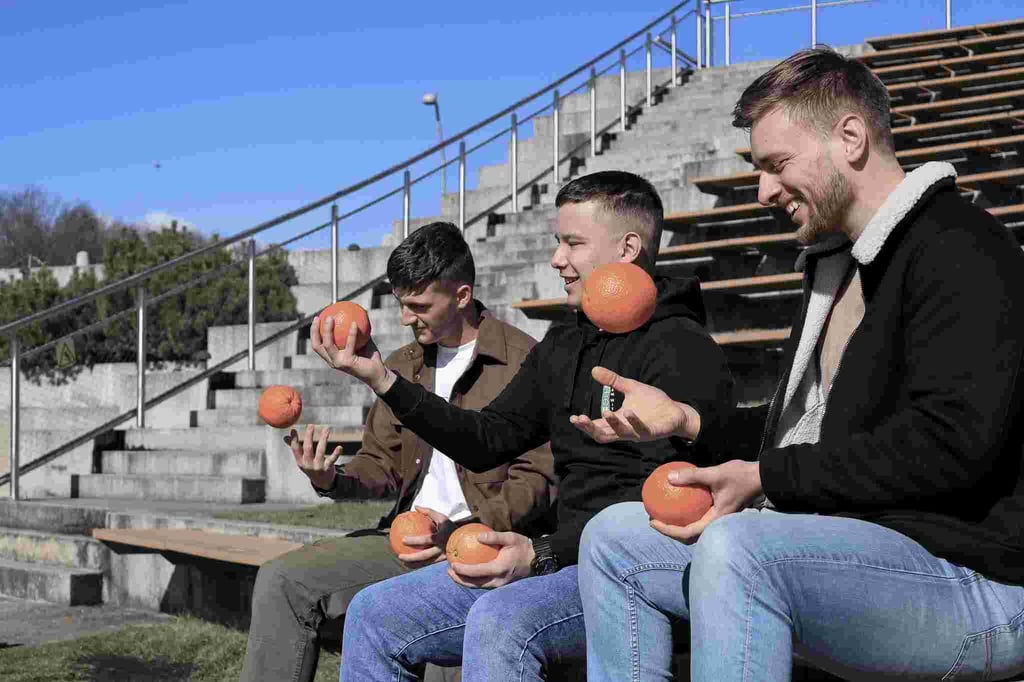 Trys jauni vyrai sedi ir meto apelsinus