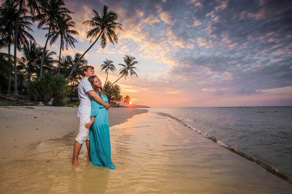 A couple embracing on a tropical beach at sunset, with palm trees and the ocean in the background