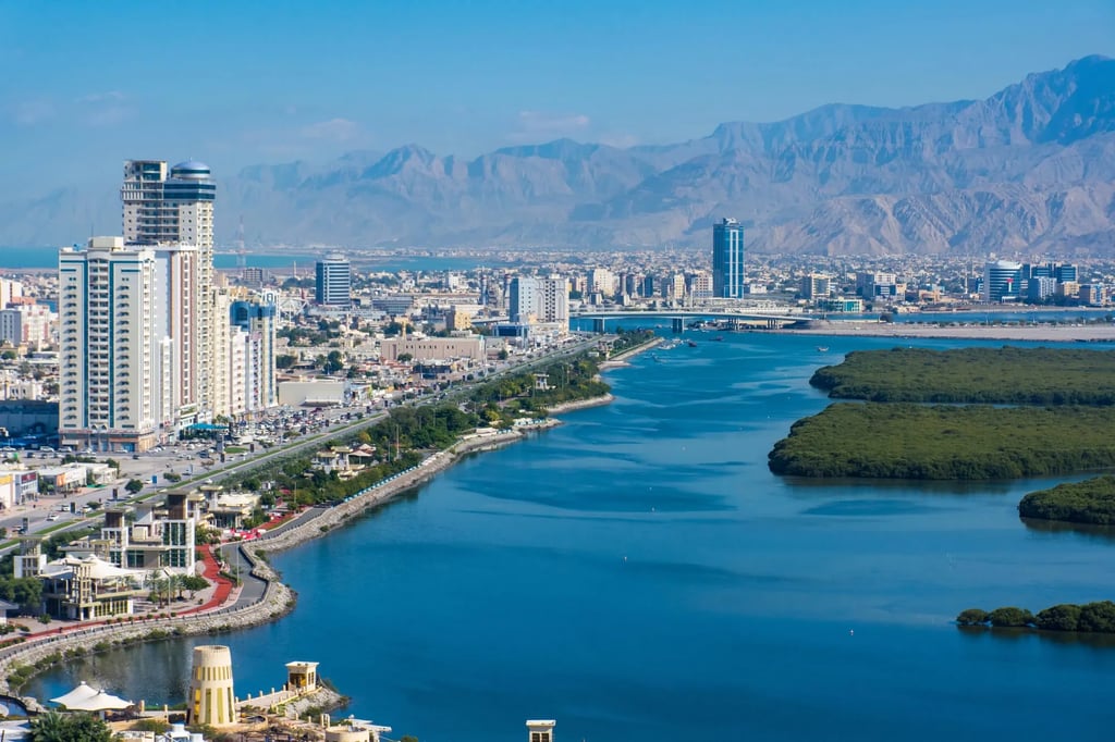 Aerial view of Ras Al Khaimah city skyline featuring skyscrapers, mangroves, and Hajar Mountains.