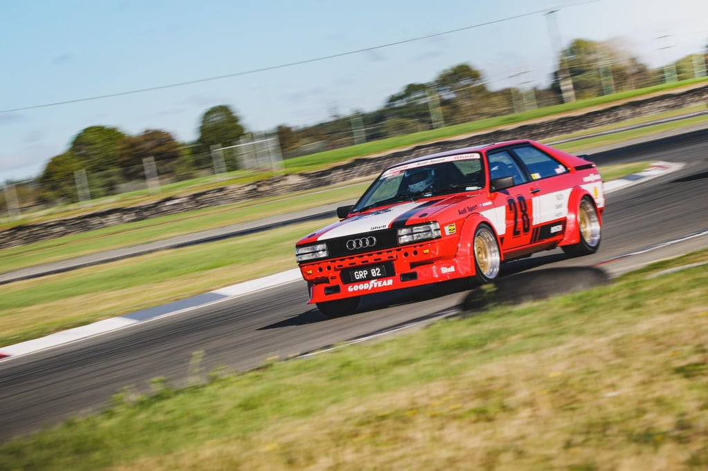 Red Audi Quattro race car with Goodyear branding speeding on a paved motorsport track.