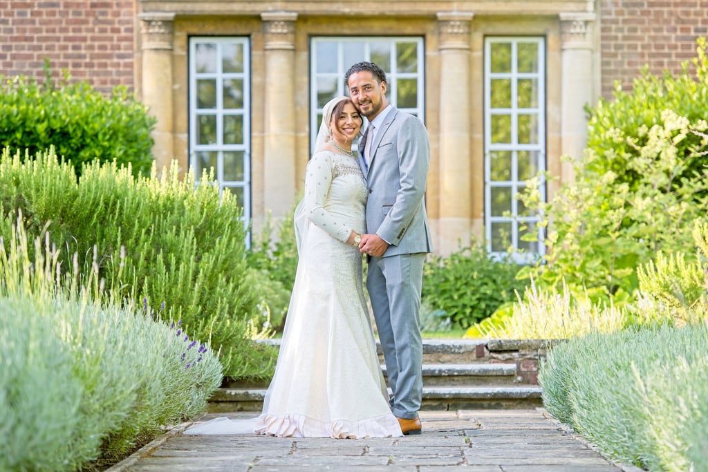 asian bride and groom holding hands and looking at camera I'm front of a Stately home, bushes are leading lines 
