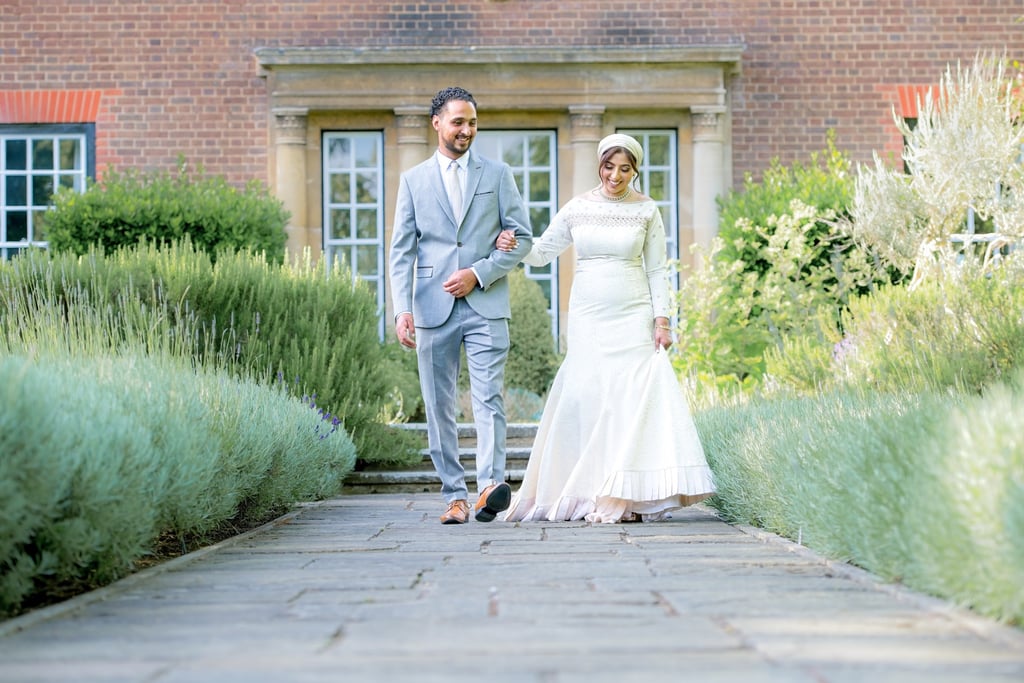 asian bride & groom outside country house walking towards camera linking arms and smiling, bushes are leading lines