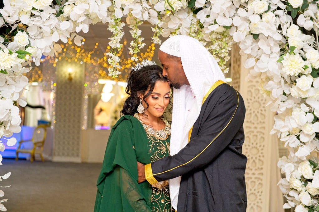 candid photo of Somalian groom kissing bride on forehead, his hugging her tight, floral arch framing them