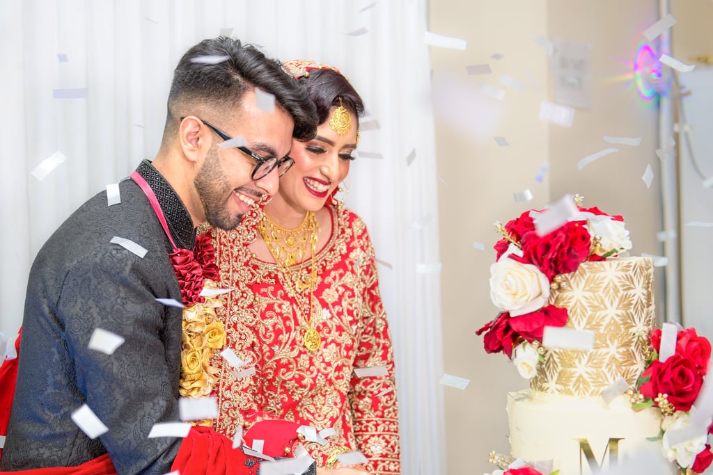 asian bride and groom in traditional outfits cutting wedding cake, both smiling, blurred confetti in foreground  
