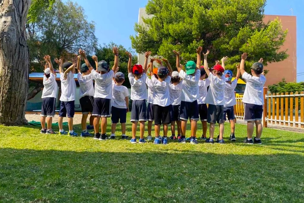 Παιδιά με καλοκαιρινές στολές και καπέλα υψώνουν τα χέρια τους στον αέρα, συμμετέχοντας σε δραστηριό