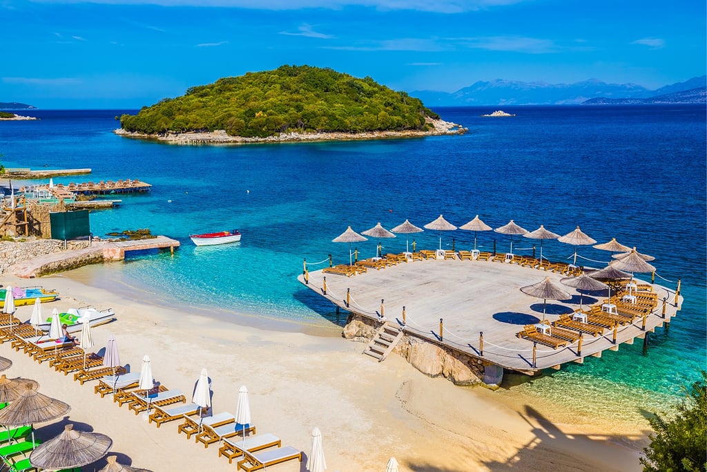 Tropical white sand beach in Ksamil, Albania, with straw umbrellas and a wooden deck overlooking turquoise waters.