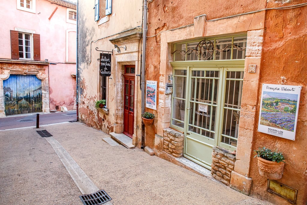 A charming narrow street in Roussillon, France, featuring terracotta buildings and an art gallery storefront.