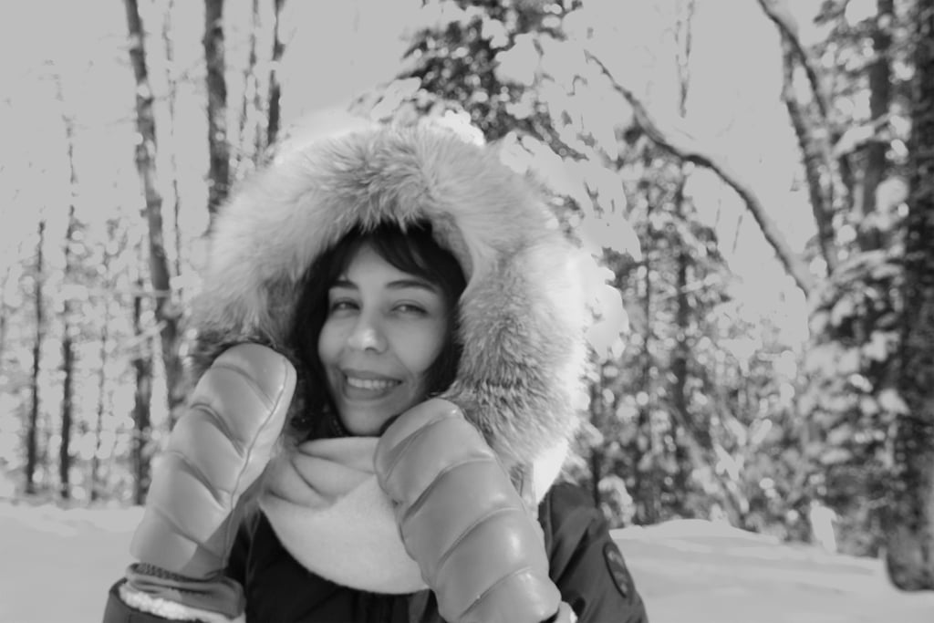 Person smiling in a snowy forest wearing a fur-lined hood and mittens.