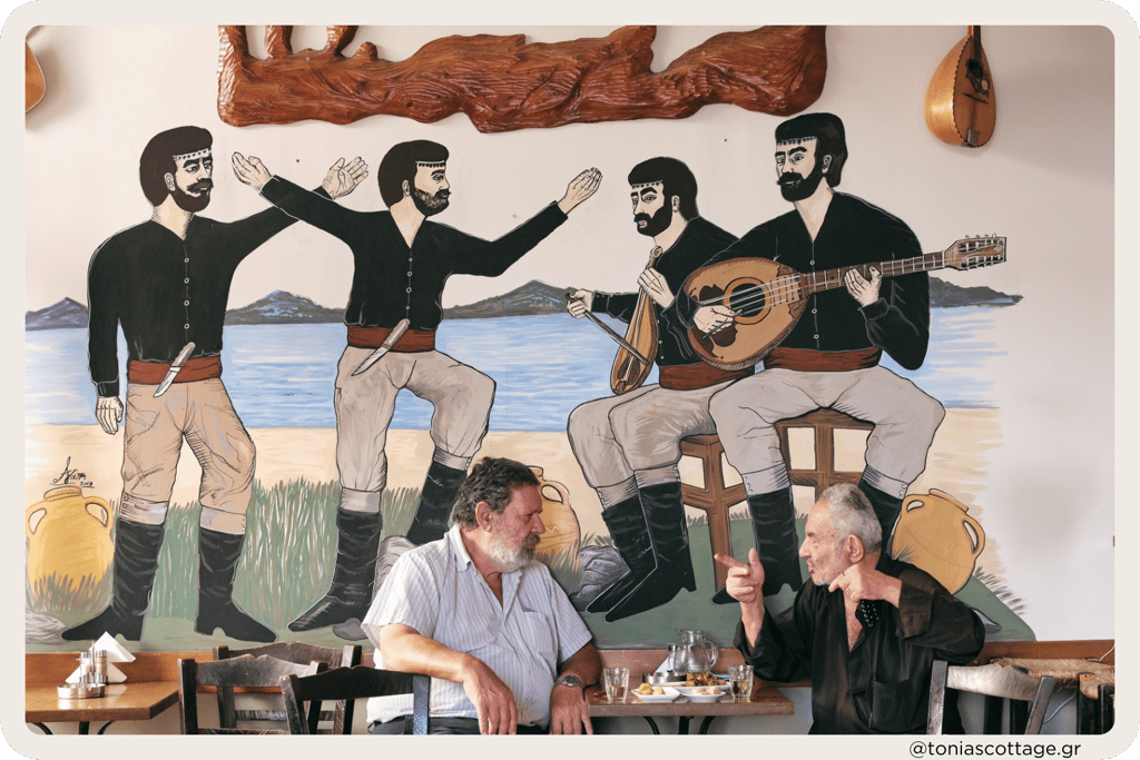 Two men sitting in a kafenio, talking, drinking coffee, with Cretan dancers painted on the wall