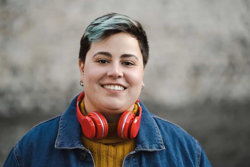 Smiling person with short blue-tipped hair wearing red headphones and a denim jacket.