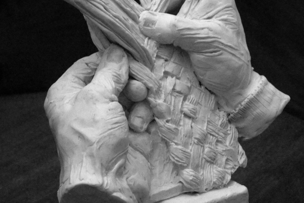 A detailed clay sculpture of two hands skillfully weaving a basket with long fibers on a square base.
