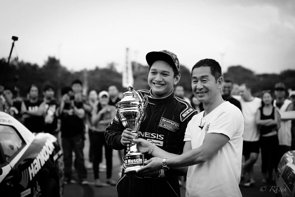 Keiichi Tsuchiya handing a trophy to a man in front of a crowd 