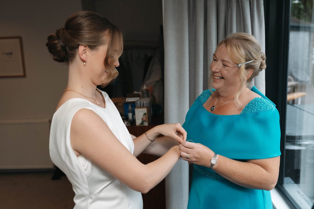 Smiling mother of the bride helps her daughter fasten a bracelet while getting ready for a wedding.