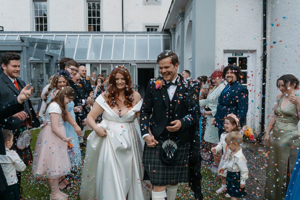 A smiling bride and groom in a traditional kilt walking through colorful wedding confetti outdoors.