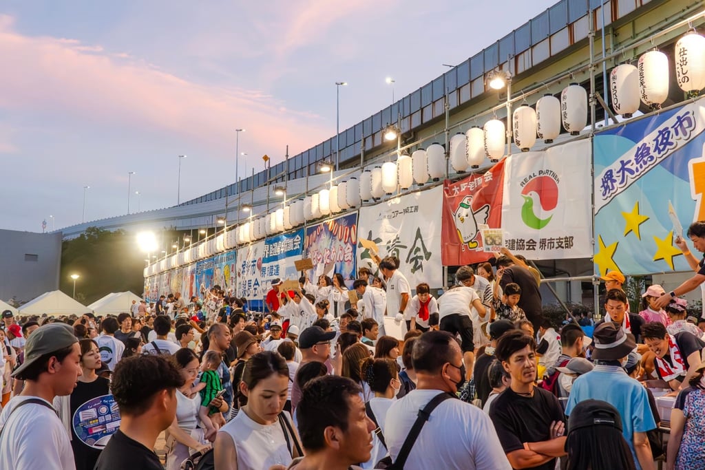 堺大魚夜市の会場で多くの来場者が魚セリを楽しむ様子。提灯と協賛団体の横断幕が並び、夏祭りのにぎわいが伝わる写真。
