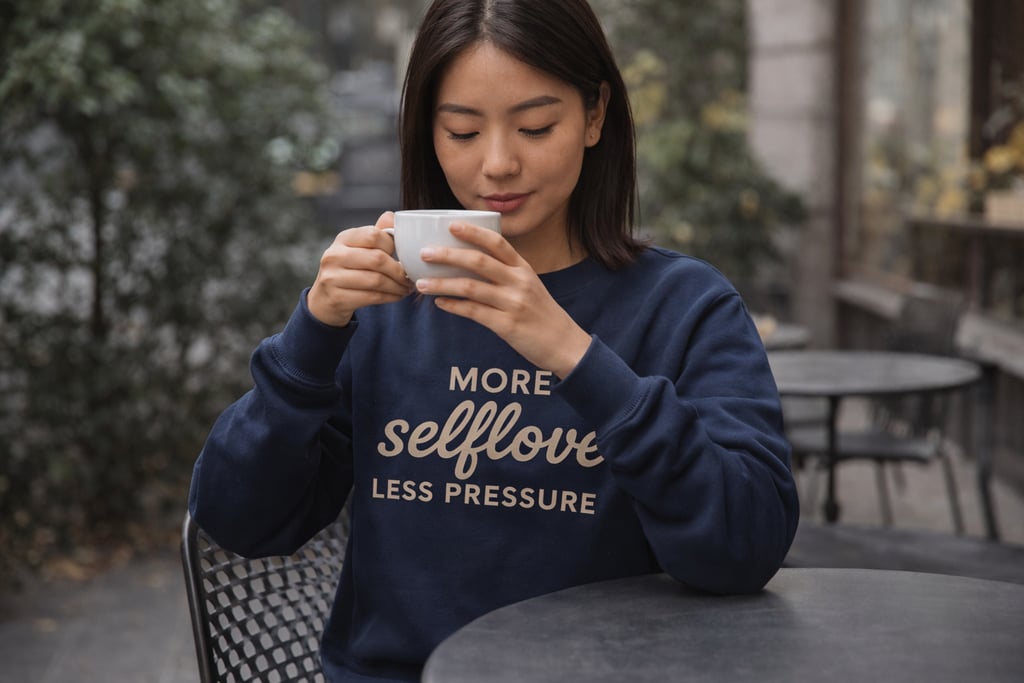 Frau mit navy Sweatshirt „More selflove less pressure“ sitzt entspannt mit Kaffee im Café