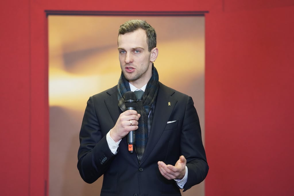 Vīrietis uzvalkā ar šalli runā mikrofonā, stāvot pie sarkanas instalācijas ar durvju atvērumu