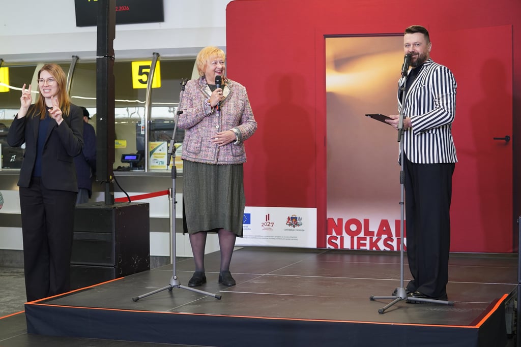 Sieviete runā mikrofonā uz skatuves pie instalācijas Nolaid slieksni, blakus vīrietis un sieviete 