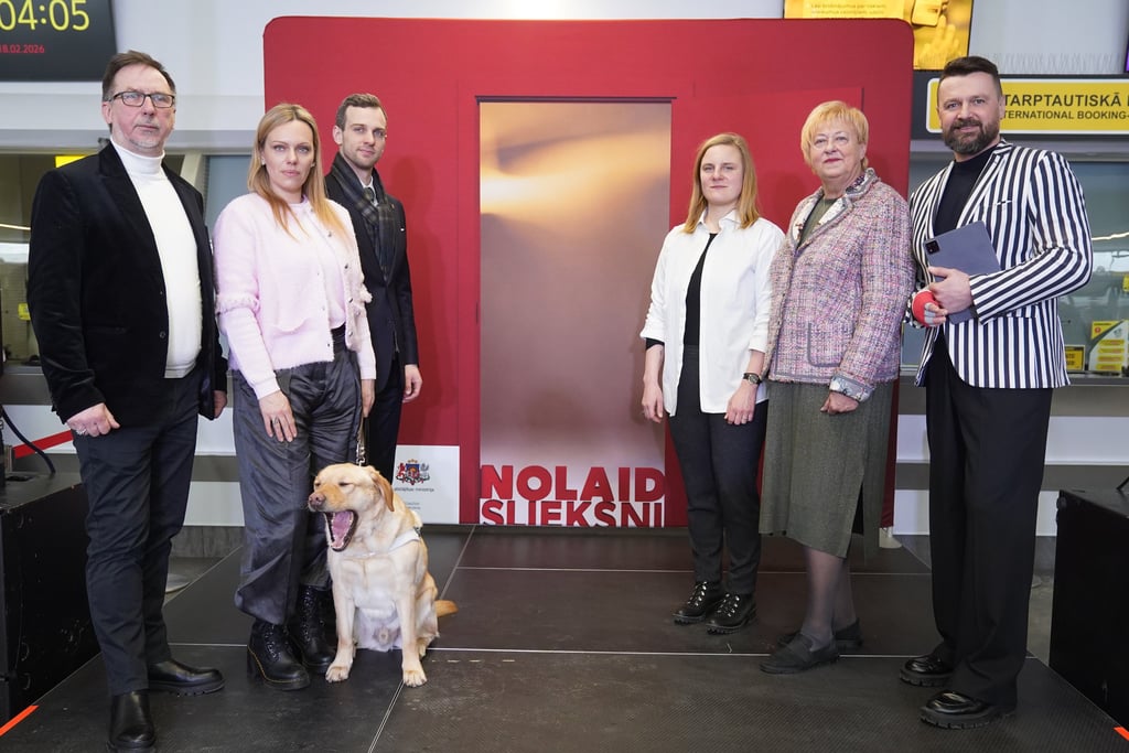 Seši cilvēki stāv pie instalācijas ar uzrakstu “Nolaid slieksni”, priekšplānā sēž suns-pavadonis