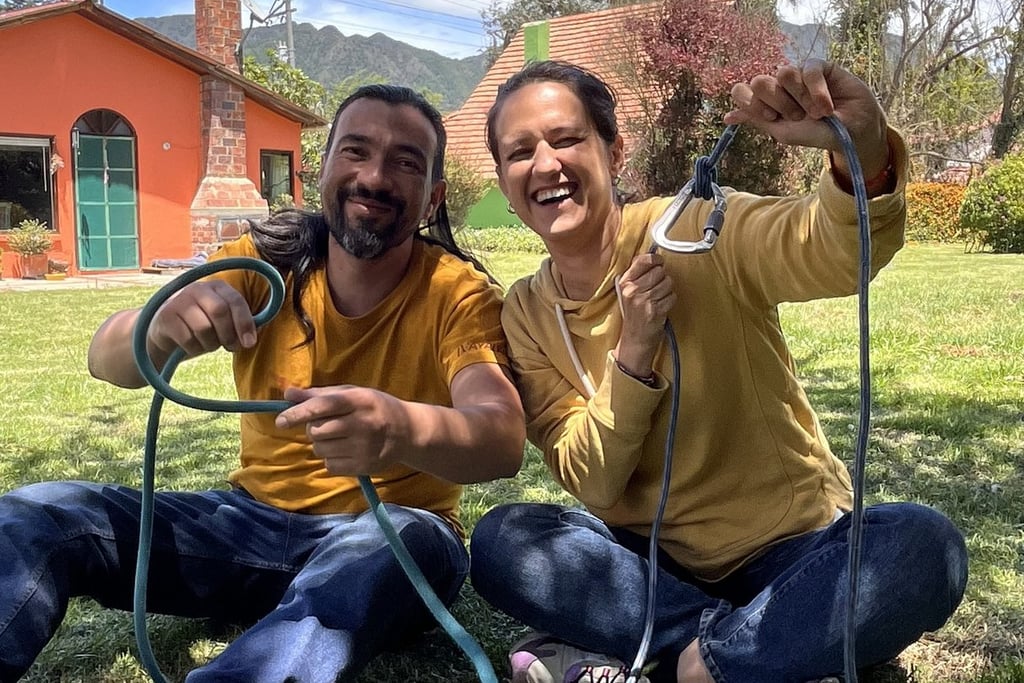 A smiling couple sits on a grassy lawn practicing rock climbing knots with a blue rope and carabiner.