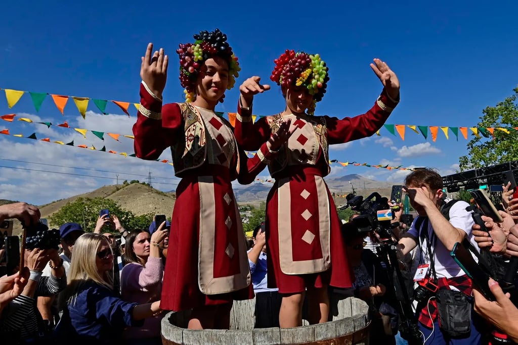Women in traditional Armenian dress wearing grape headpieces crush grapes at a harvest festival.