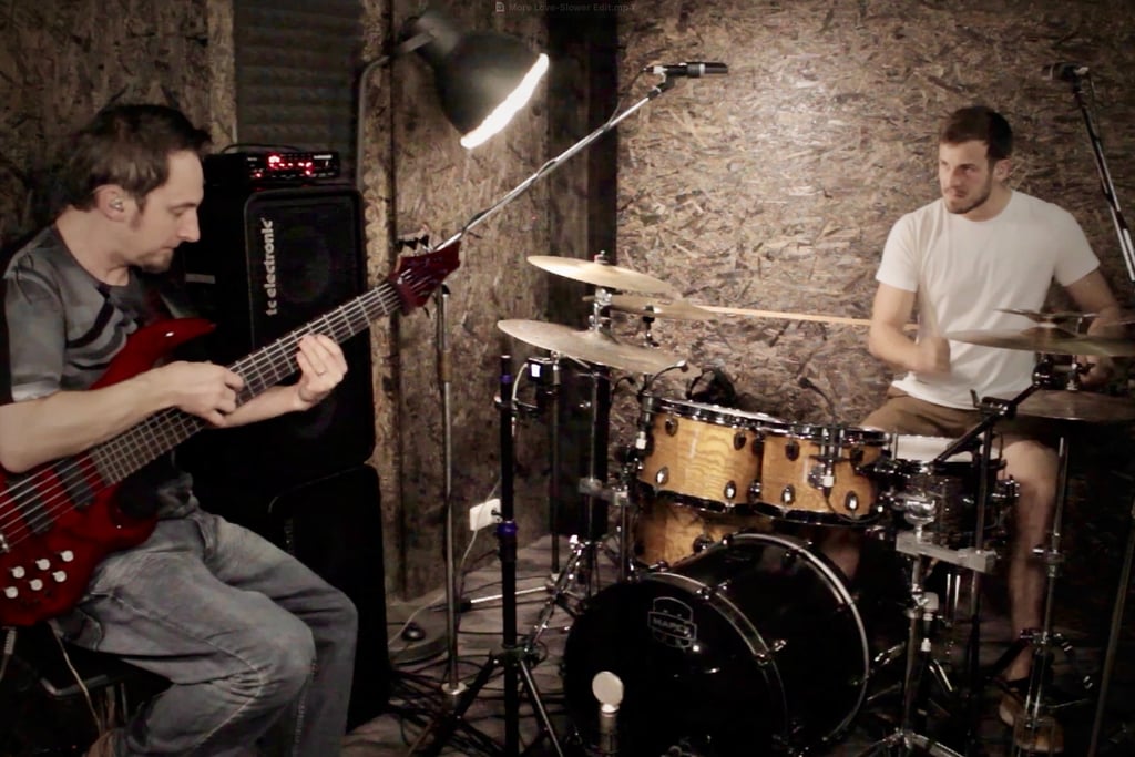 Jozef Bobula on red six-string bass with drummer in a rustic dimly lit rehearsal space.