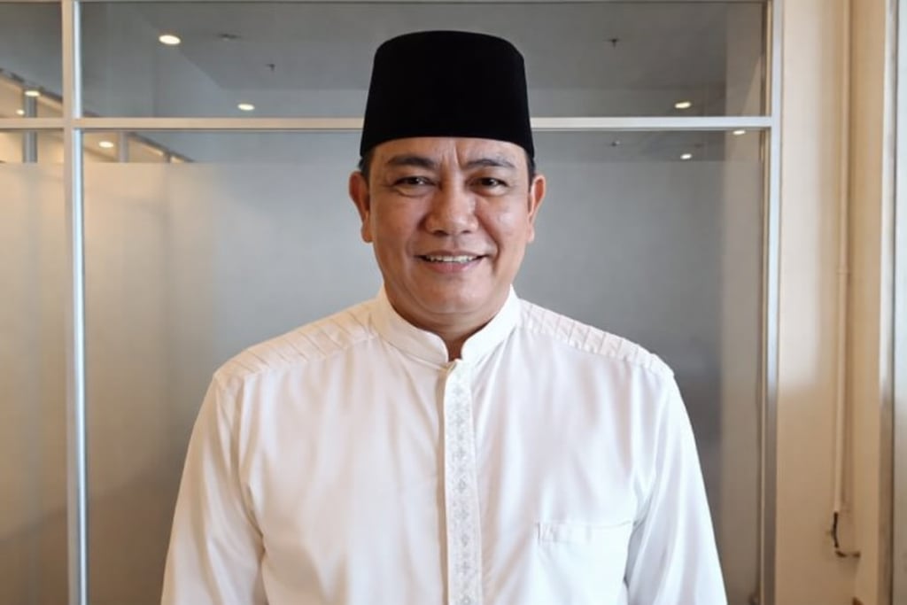 Smiling man wearing a traditional black songkok hat and white baju melayu shirt in an office setting.
