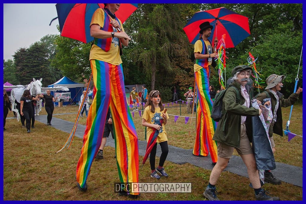 Performers on stilts wearing rainbow trousers and holding umbrellas at an outdoor festival parade.