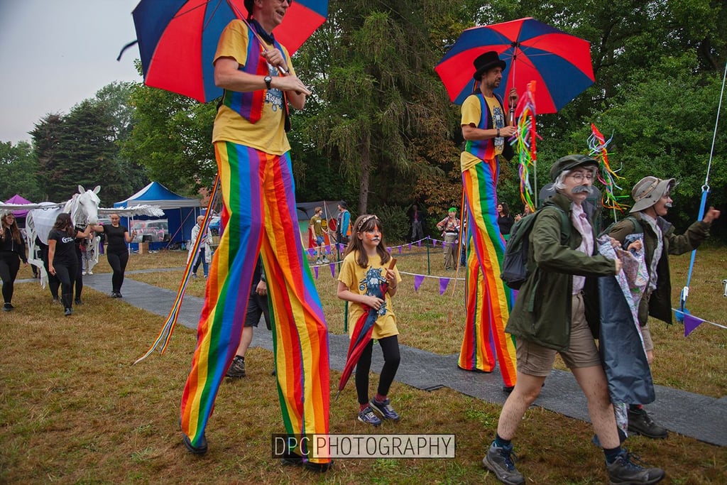 Performers on stilts wearing rainbow trousers and holding umbrellas lead a parade at a festival.