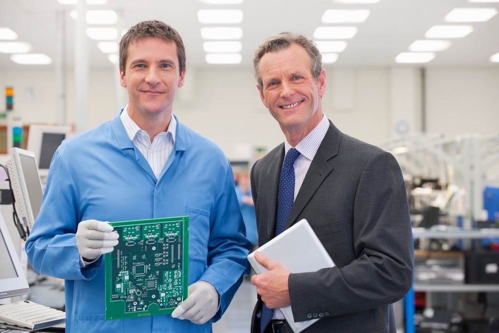 Engineer and business partner reviewing electronic circuit board in production facility.