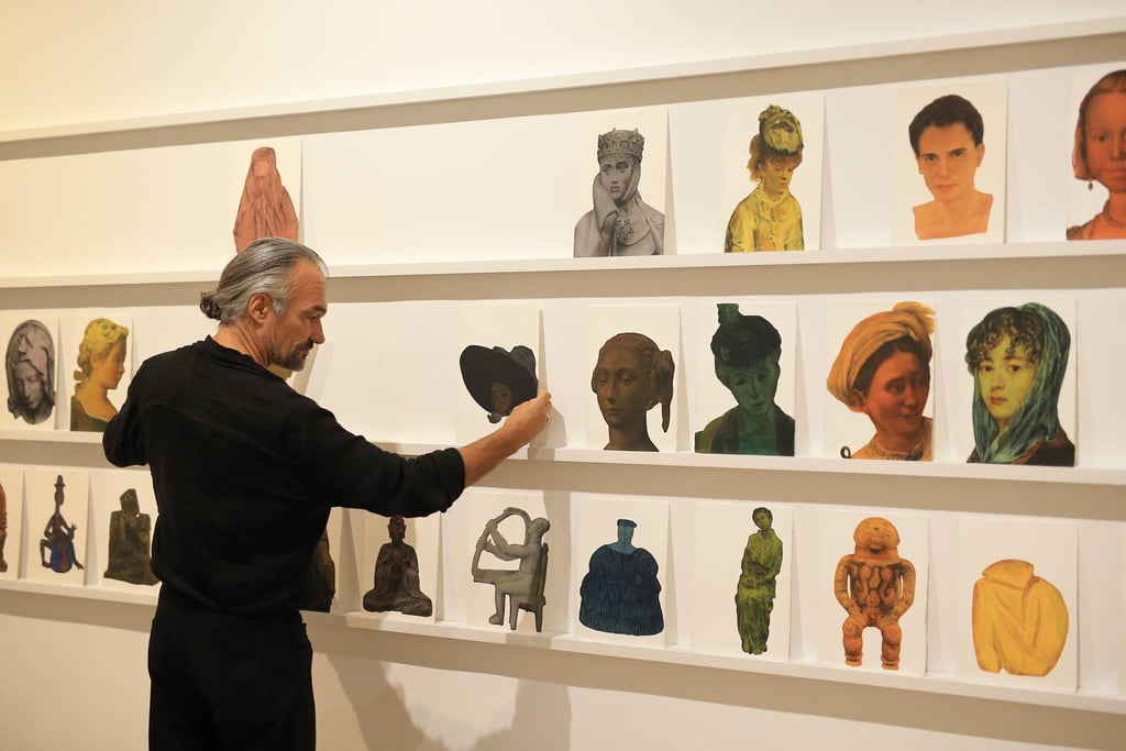 An artist arranges a diverse collection of portrait paintings on white gallery shelves for an exhibition.