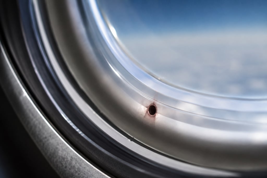 Close-up of a small breather hole in an airplane window overlooking clouds.